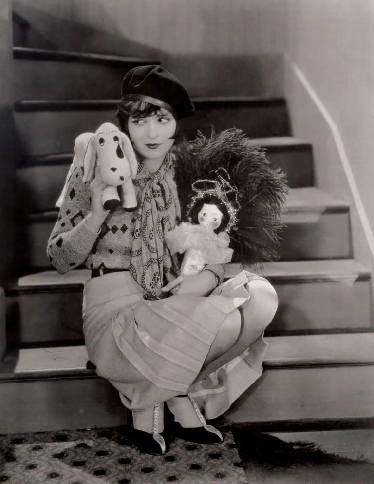 Black-and-white 1920s studio photo: a young woman in a beret, patterned sweater and pleated skirt crouches on a staircase holding a toy dog, a doll and a feather fan