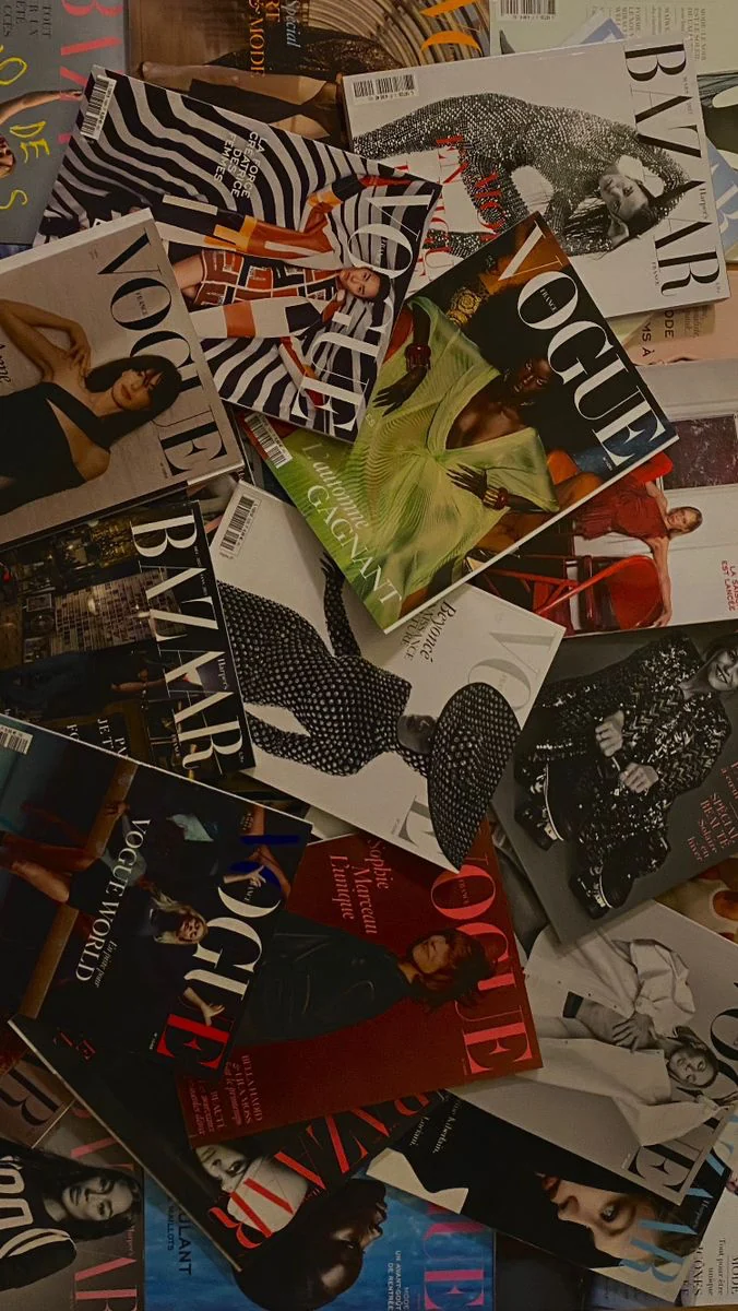 A pile of printed fashion magazines, Vogue and Harper’s Bazaar covers in French and English, scattered on top of each other in warm, dim light