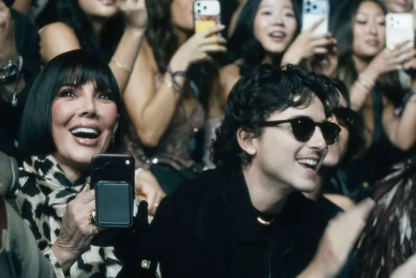 Timothée Chalamet in sunglasses and Kris Jenner smiling in the front row, surrounded by guests filming on their phones